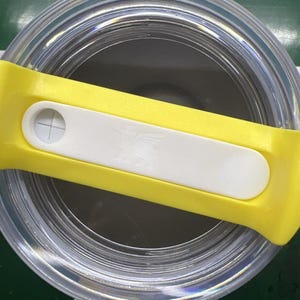 May include: A clear, cylindrical container with a yellow and white lid. The lid has a white rectangular button with a cross symbol. The container appears to be made of clear plastic or glass.