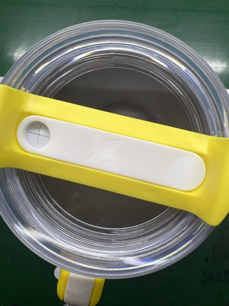 May include: A clear, cylindrical container with a yellow and white lid. The lid has a white rectangular button with a cross symbol. The container appears to be made of clear plastic or glass.