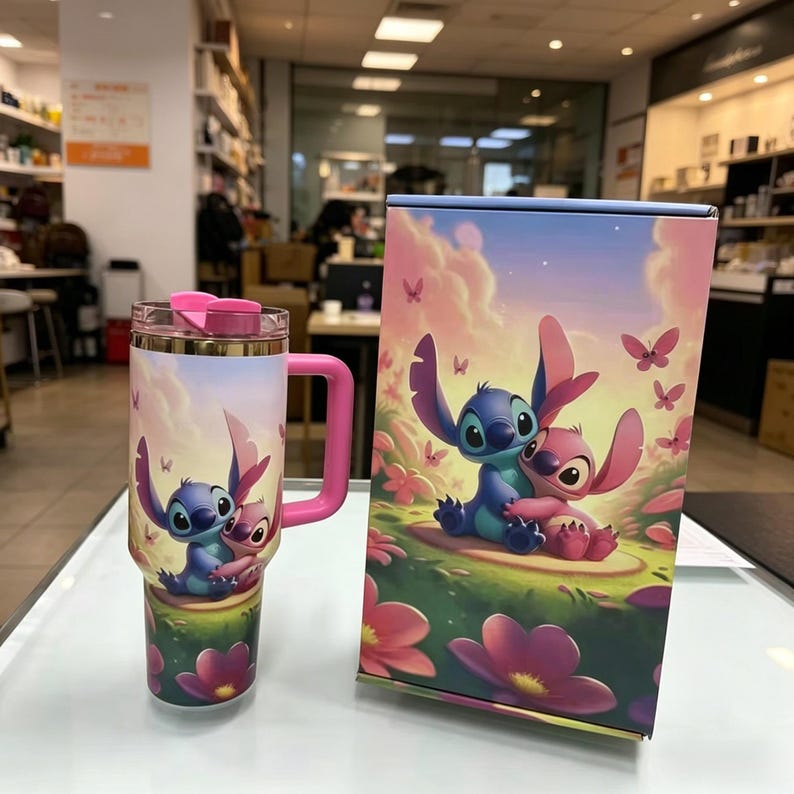 May include: A stainless steel tumbler with a pink handle and a clear lid featuring a pink flip-top, decorated with a graphic of Stitch and Angel hugging in a field of flowers. Beside it stands a rectangular box displaying the same character artwork.