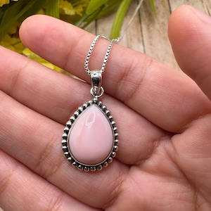 May include: A silver pendant necklace featuring a teardrop-shaped, light pink gemstone. The gemstone is surrounded by a beaded silver border and hangs from a delicate silver chain. The necklace is displayed on a wooden surface.