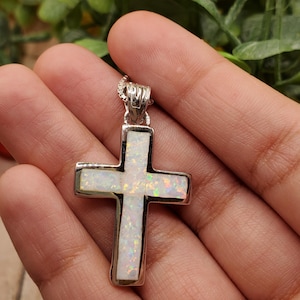 May include: A silver-toned pendant featuring a cross design. The cross is filled with a white, iridescent material, and has a silver border. The pendant is held in a person's hand, with a blurred green background.