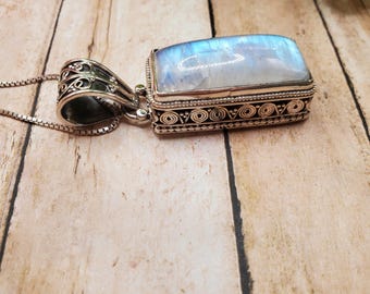 May include: A rectangular pendant with a large, iridescent, blue-white stone set in a silver-colored metal frame. The pendant hangs from a delicate chain. The frame has an ornate, detailed design. The background is a light brown wooden surface.