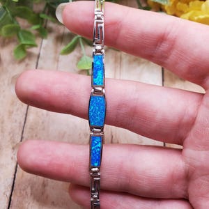 May include: A silver-toned link bracelet featuring three rectangular inlays of vibrant blue opal-like material. The bracelet is constructed with alternating metal links that display a geometric Greek key pattern between the blue stone segments.