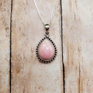 May include: A teardrop-shaped pink stone pendant set in a sterling silver frame with a beaded edge. The pendant hangs from a thin silver box chain necklace. The stone has a smooth, polished surface with subtle variations in its light pink colour.