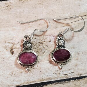 May include: A pair of silver-tone dangle earrings. Each earring features an oval, deep pink gemstone set in a silver frame. The earrings have a decorative top with a small, round accent stone and a hook closure.