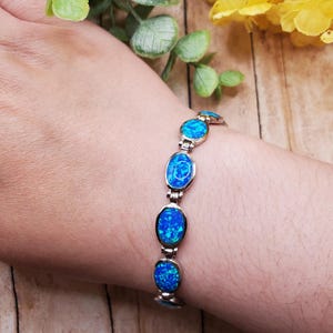 May include: A silver-toned link bracelet featuring a series of oval-shaped, vibrant blue iridescent stones. The bracelet is constructed with metal hinges connecting each individual stone setting, creating a flexible chain design that rests against the wrist.