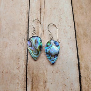 May include: A pair of teardrop-shaped earrings featuring iridescent abalone shell inlays set in polished silver frames. Each earring hangs from a curved silver ear wire, showcasing swirling patterns of blue, green, and purple hues within the shell.