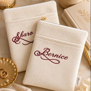 May include: Two cream-colored corduroy pouches with gold-tone zippers. Each pouch features custom embroidery in dark red script; one reads Sharo and the other reads Bernice. The pouches have a horizontal stitched seam across the upper section.