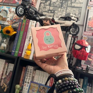 May include: A pink box with the text "by Kenny" and a cartoon lock design. The box is held in a hand adorned with multiple bracelets. The background includes bookshelves, comic books, and a toy motorcycle.