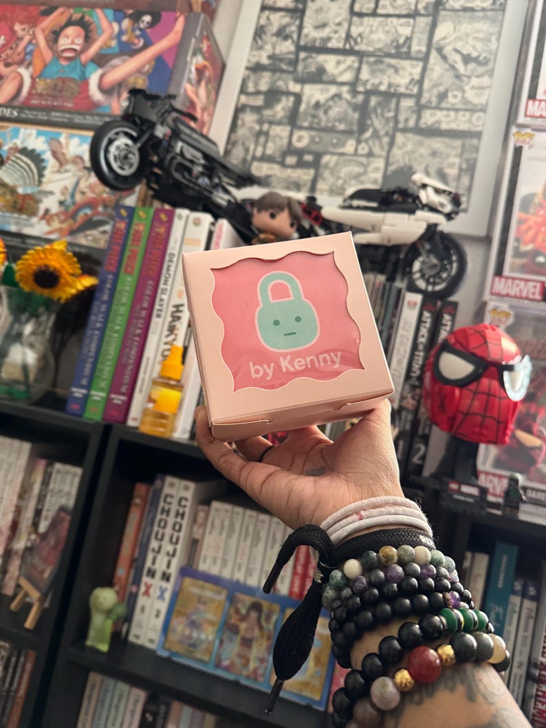 May include: A pink box with the text "by Kenny" and a cartoon lock design. The box is held in a hand adorned with multiple bracelets. The background includes bookshelves, comic books, and a toy motorcycle.