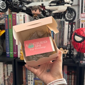 May include: A person holding a light pink box with the text "LOCKED IN by Kenny" on a pink card. The box is open, revealing a wrapped item. The background includes bookshelves, toy motorcycles, and figurines.