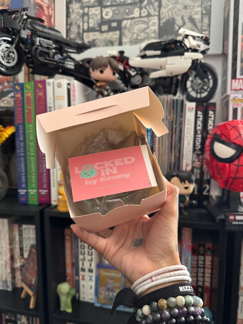 May include: A person holding a light pink box with the text "LOCKED IN by Kenny" on a pink card. The box is open, revealing a wrapped item. The background includes bookshelves, toy motorcycles, and figurines.