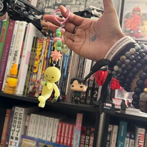 May include: A handmade keychain featuring a yellow figure with a green accessory, suspended from a pink heart-shaped clasp. The keychain is adorned with pastel-colored beads and charms. The background includes bookshelves filled with books and collectibles.