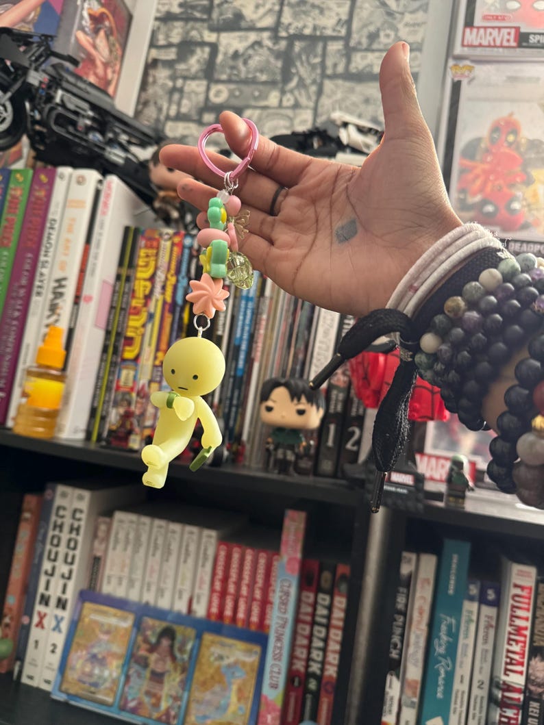 May include: A handmade keychain featuring a yellow figure with a green accessory, suspended from a pink heart-shaped clasp. The keychain is adorned with pastel-colored beads and charms. The background includes bookshelves filled with books and collectibles.