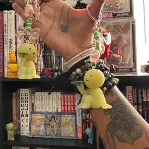 May include: A pair of keychains with yellow, cartoon-like figures. One keychain has a green bow and the other has a tray. Both keychains have pink keyrings and are adorned with pink, green, and clear beads. The background includes books and comic book cases.