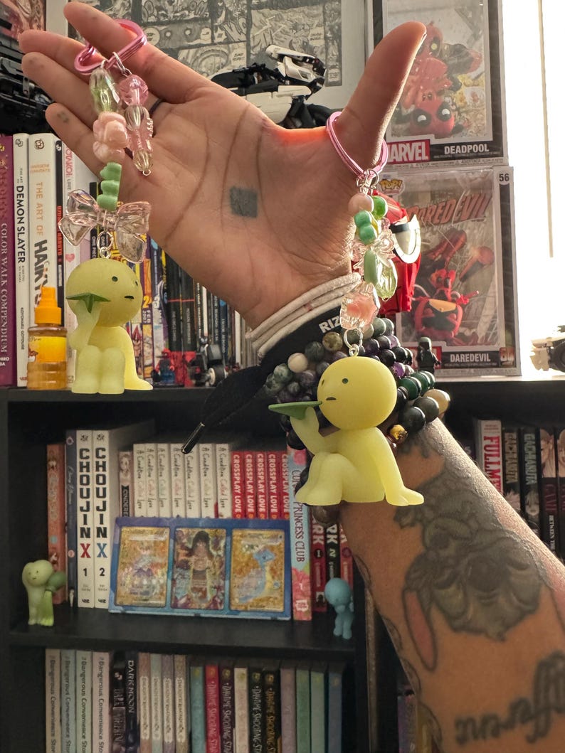 May include: A pair of keychains with yellow, cartoon-like figures. One keychain has a green bow and the other has a tray. Both keychains have pink keyrings and are adorned with pink, green, and clear beads. The background includes books and comic book cases.