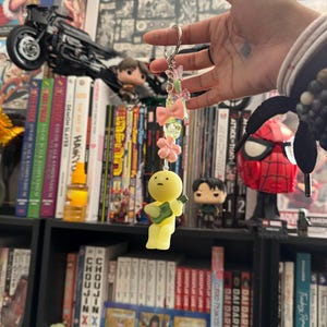 May include: A keychain with a yellow figure holding a guitar, adorned with pink bows, flowers, and beads. The keychain is suspended in front of a bookshelf filled with manga and comic books, including "Demon Slayer" and "Attack on Titan."