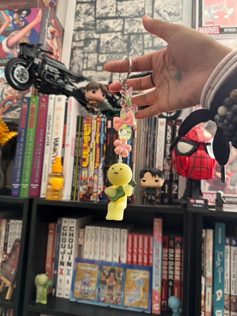 May include: A keychain with a yellow figure holding a guitar, adorned with pink bows, flowers, and beads. The keychain is suspended in front of a bookshelf filled with manga and comic books, including "Demon Slayer" and "Attack on Titan."