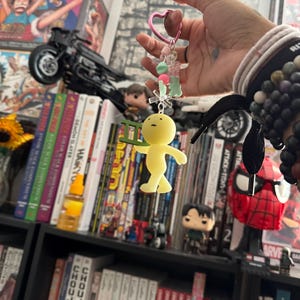 May include: A yellow figurine keychain with a ladder, held by a pink heart-shaped clasp. The keychain has green and pink beads. The background includes a bookshelf with manga and figurines.