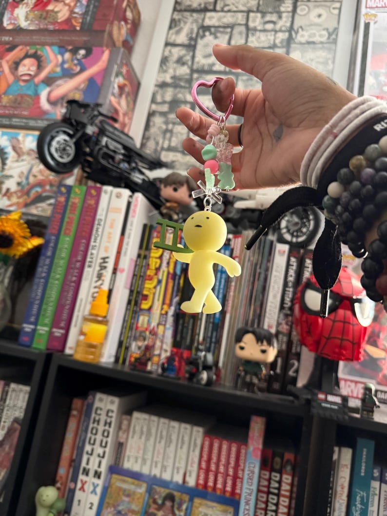 May include: A yellow figurine keychain with a ladder, held by a pink heart-shaped clasp. The keychain has green and pink beads. The background includes a bookshelf with manga and figurines.