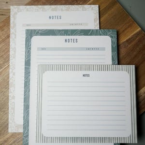 May include: A stack of notepads with the word "NOTES" at the top. The notepads have different designs, including floral patterns and striped borders. The notepads are white, gray, and teal, and are arranged on a wooden surface.