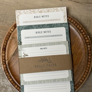 May include: A stack of three rectangular paper notepads labeled BIBLE NOTES, held together by a brown paper band printed with HELLO FAITH CO. The top pad has a beige floral border, the middle has a green botanical print, and the bottom has green stripes.