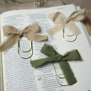 May include: Three decorative bow-shaped page clips with gold-colored metal clasps. The bows are made of textured fabric in beige, olive green, and tan colors. The clips are resting on an open book.