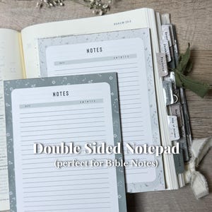 May include: A double-sided notepad with the word "NOTES" at the top, featuring a floral border design. The notepad is shown next to an open book with labeled tabs. The text "Double Sided Notepad" is displayed on the notepad.