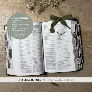 May include: An open Bible with dark green covers, featuring numerous labeled tabs along the edges. A dark green ribbon bow is tied to the top of the pages. A circular overlay lists included items: notepad, pen, Bible book tracker, 3 ribbon paperclips, and 3 button pins.
