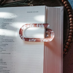 Puede incluir: Un sujetapáginas transparente en forma de C con un diseño floral en tonos rosa y blanco, apoyado en un libro abierto. El texto "PSALM 119:138" es visible en la página. El libro está abierto en una página de texto.