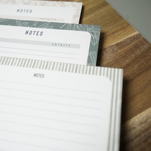 May include: Close-up of three notepads with the word "NOTES" printed on the top. The notepads have lined pages and floral or striped designs. They are resting on a wooden cutting board and a gray surface.