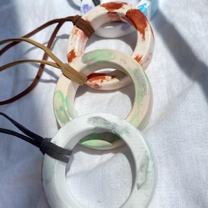 May include: Four circular pendants on leather cords. The top pendant is white with blue and purple splotches. The second is white with rust-colored markings. The third is white with green and orange accents. The bottom pendant is white with gray marbling.