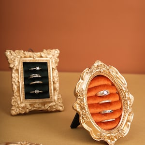 May include: Ornate gold-colored ring display frames. One frame is rectangular with a dark green velvet interior, the other is oval with an orange velvet interior. Several rings are displayed in each frame, showcasing jewelry.