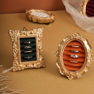 May include: Ornate gold-colored ring displays. One rectangular frame holds rings against a dark green backdrop, while an oval frame displays rings against an orange backdrop. Additional gold frames are in the background.