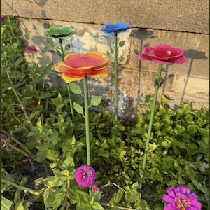 May include: A garden scene features colorful metal flower sculptures in red, yellow, blue, and green. The flowers are on green stems with leaves, set among green foliage and other pink flowers. The background includes a brick wall.