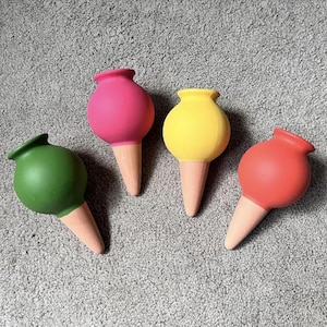 May include: Four colorful, decorative bottle stoppers shaped like ice cream cones. Each stopper has a round, brightly colored top in green, pink, yellow, and red, with a light brown cone base. The stoppers are arranged on a gray surface.
