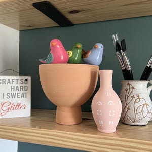May include: A wooden shelf displays a variety of decorative items. There are three ceramic bird figurines, a large peach-colored bowl, a pink vase with white accents, and a ceramic mug with paintbrushes. A small sign reads "I Craft So Hard I Sweat Glitter."