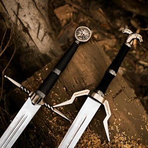 May include: Two silver and black swords with intricate designs. One sword features a wolf emblem on the pommel, while the other has a bird-like design. Both have black wrapped handles and silver crossguards, resting on a wooden surface.