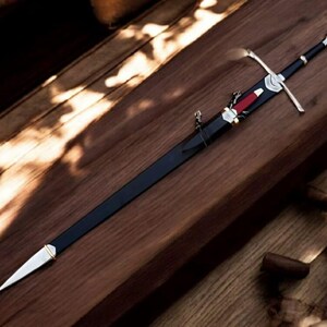 May include: A long, black sword with a silver crossguard and a red accent on the sheath. The sword is resting on a wooden surface, showcasing its detailed craftsmanship and design. The blade is sheathed in black.