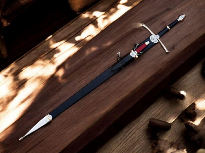 May include: A long, black sword with a silver crossguard and a red accent on the sheath. The sword is resting on a wooden surface, showcasing its detailed craftsmanship and design. The blade is sheathed in black.