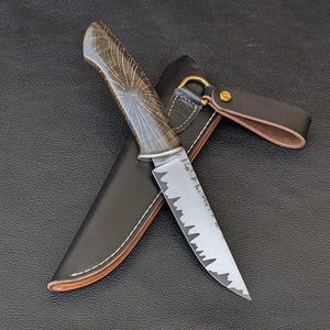Könnte beinhalten: Ein handgefertigtes Jagdmesser mit gemustertem Holzgriff und gezahnter Klinge. Das Messer liegt in einer schwarzen Lederscheide mit braunem Lederriemen und Messingbeschlägen. Die Klinge hat ein einzigartiges Walddesign.