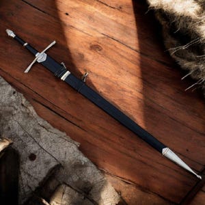 May include: A long, black sword with a silver crossguard and accents, lying on a wooden surface. The sword has a pointed tip and a black handle. The image is a top-down view of the sword, with a rustic, medieval aesthetic.