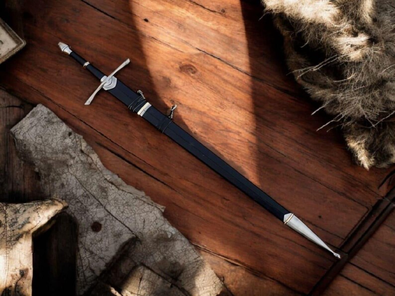 May include: A long, black sword with a silver crossguard and accents, lying on a wooden surface. The sword has a pointed tip and a black handle. The image is a top-down view of the sword, with a rustic, medieval aesthetic.