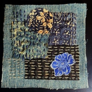 May include: A square denim fabric patch featuring visible sashiko-style stitching in white and dark blue. The design includes a circular tan floral fabric appliqué, a dark blue geometric patterned square, and a bright blue floral fabric appliqué.