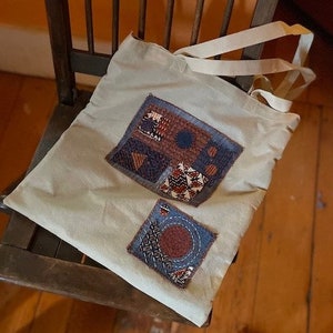 May include: A white canvas tote bag with two colorful fabric patches sewn on. The patches are made of blue, brown, and orange fabric and have a geometric pattern.