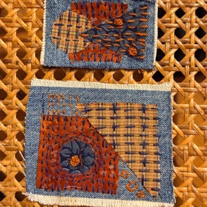 May include: Two denim patches with intricate embroidery. The patches feature a mix of orange, blue, and tan fabrics, with detailed stitching creating geometric and floral patterns. The edges of the denim are frayed.