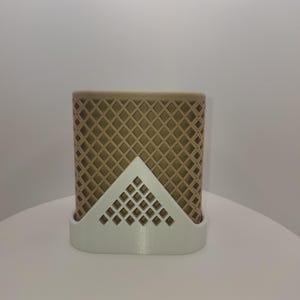 Può includere: Un contenitore rettangolare beige con un motivo a rombi, tenuto da un supporto triangolare bianco. Il supporto ha un design geometrico. Il contenitore è probabilmente per contenere oggetti.
