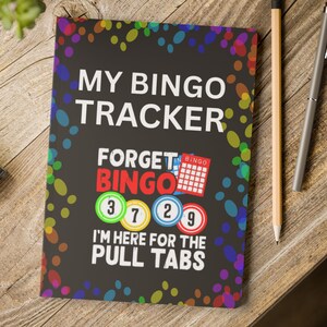 Può includere: Un taccuino nero per il bingo con il testo "MY BINGO TRACKER" e "FORGET BINGO". Il taccuino ha accenti colorati a forma di coriandoli e i numeri 3, 7, 2 e 9. Una matita e una penna sono accanto al taccuino.