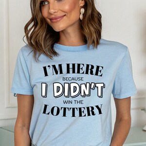 Puede incluir: Camiseta azul claro con el texto "I'M HERE BECAUSE I DIDN'T WIN THE LOTTERY" en blanco y negro. La camiseta está hecha de un material suave y tiene cuello redondo.