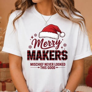 May include: A white short-sleeve t-shirt featuring a red Santa hat graphic above the text Merry MAKERS in a red and black buffalo plaid pattern, with the additional text MISCHIEF NEVER LOOKED THIS GOOD printed in red below the main design.